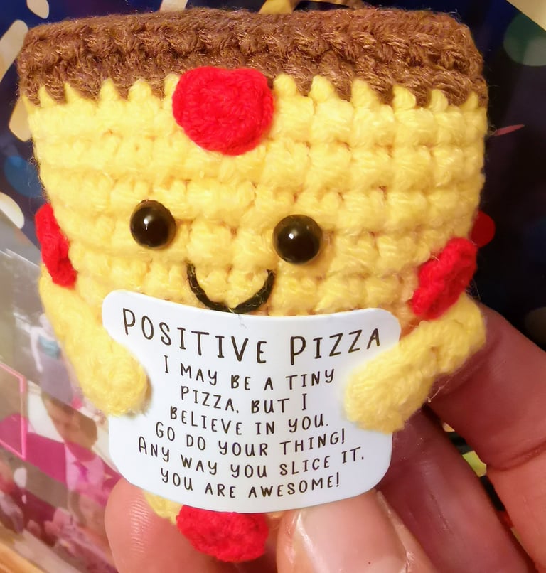 Small yellow crochet pizza amigurumi holding a positive message card with an encouraging quote.