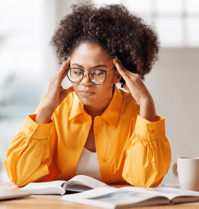 a woman in glasses and yellow shirt is sitting at a table with laptop and notepads looking stressed