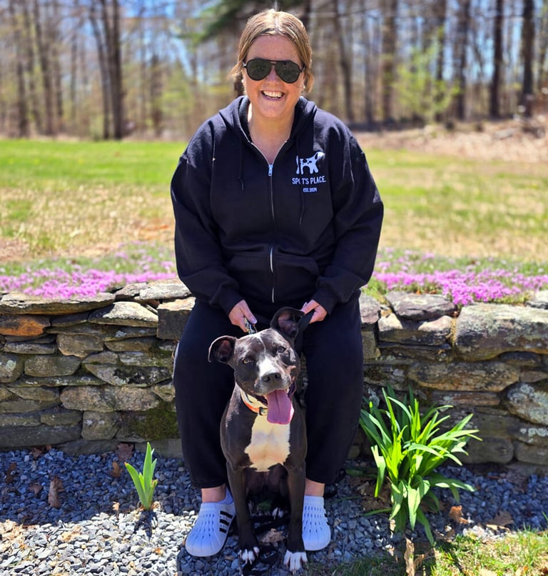 A smiling woman in sunglasses sits on a stone wall with a happy black and white dog. 