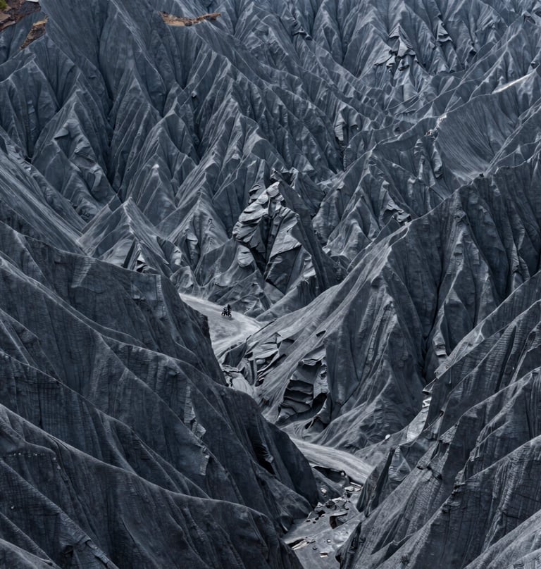 A sprawling, minimalist landscape of Global / Western canyon walls. The rock faces are shades of dark grey and cool grey. The composition is dominated by negative space, with a small, distant motorcycle visible for scale.