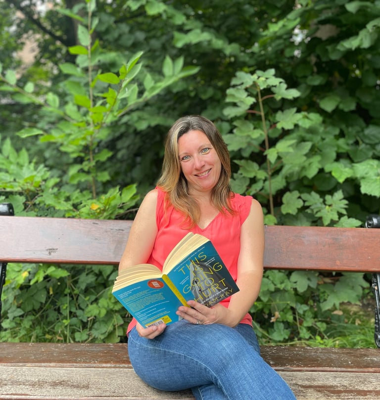 Portrait photo of Jen Ruthe sitting on a park bench in a red top, holding a book.