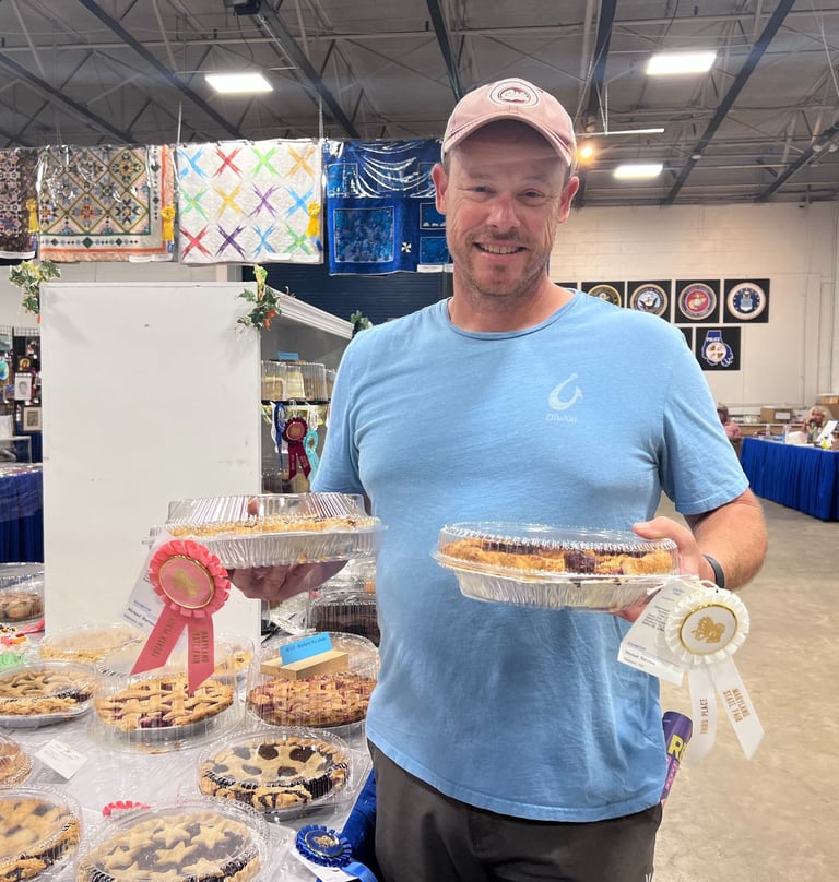 A man smiling at a county fair home arts competition holding award-winning baked fruit pies with ribbons.