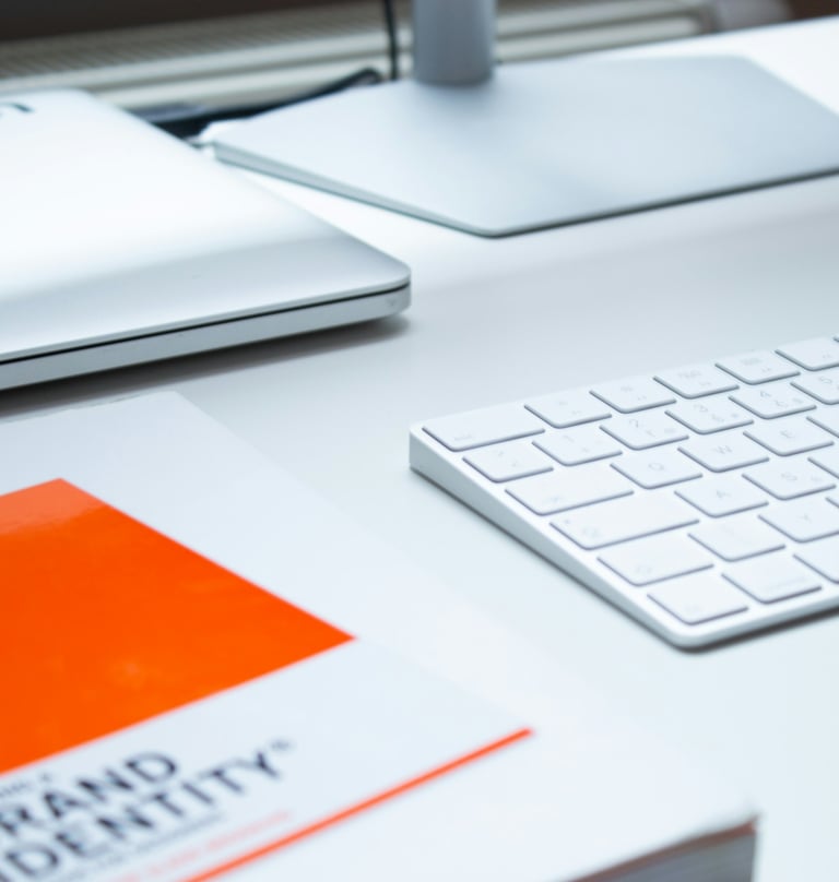 A "Brand Identity" book on a white desk with a keyboard, monitor, and laptop.