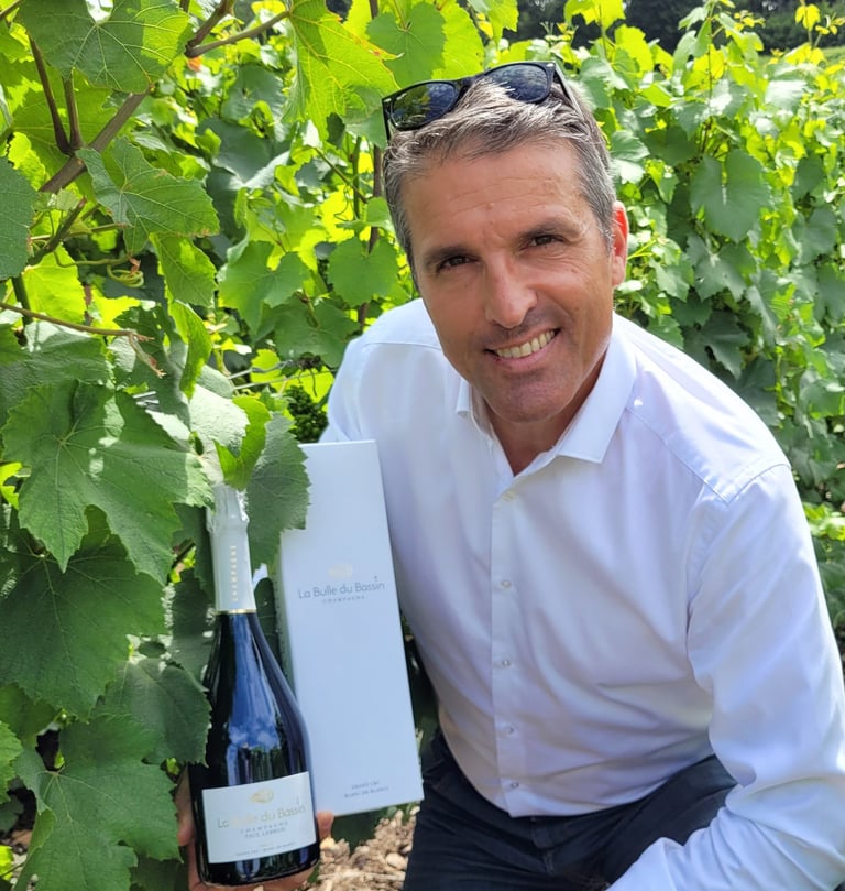 Patrice agenouillé dans les vignes tenant une bouteille de Champagne La Bulle du Bassin et son étui
