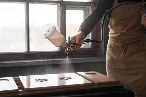 Professional woodworker using a pneumatic spray gun to apply a clear varnish finish to wooden boards.