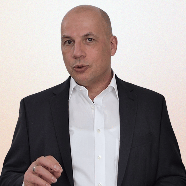 A bald male professional wearing a black suit jacket and white shirt speaking during a presentation.