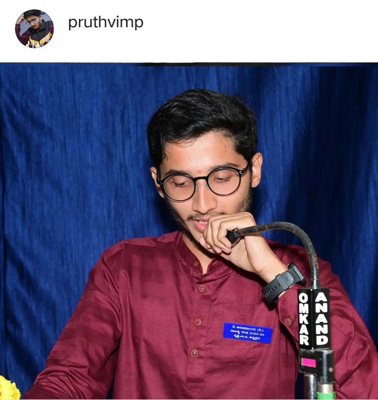 Pruthvi M P in a red shirt and glasses is holding a microphone