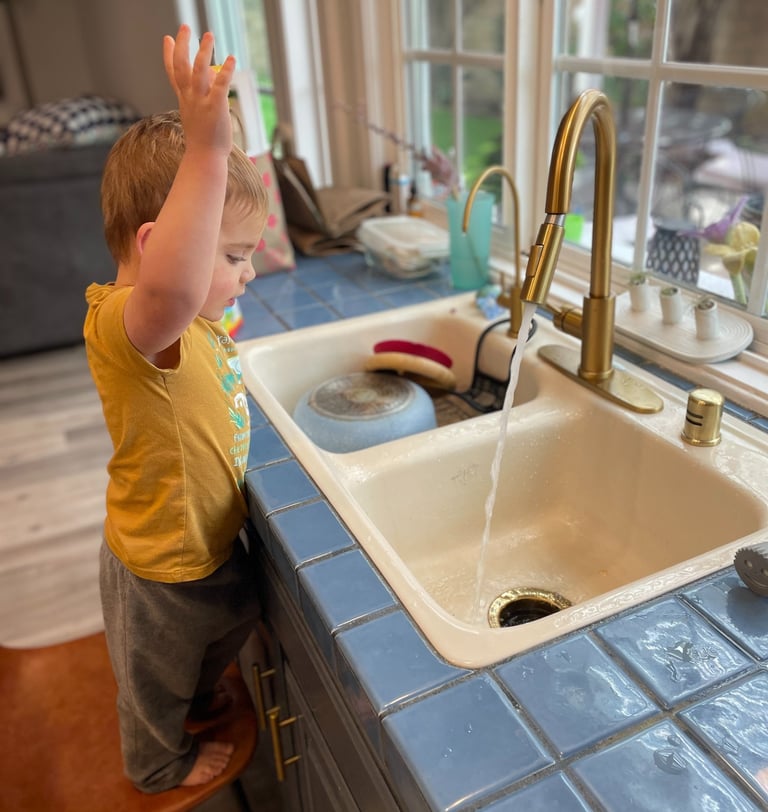 little boy standing on chair at kitchen sink with hands in the air