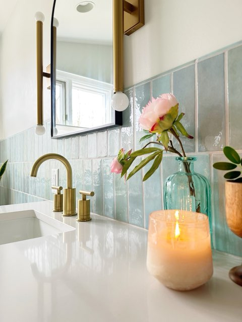Modern bathroom vanity with light blue subway tile, gold faucet, and a lit candle.