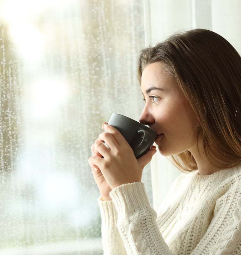 a woman drinking from a mug sitting by a window and its raining outisde