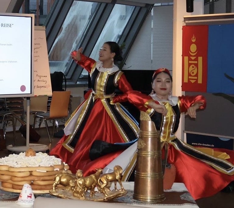 Mongolian dancers in traditional red deel costumes performing at a cultural event with folk decor.