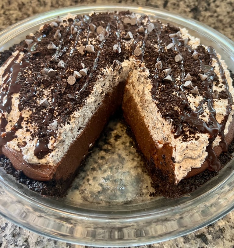 A homemade chocolate silk pie with a sliced section, topped with whipped cream, cookie crumbles, and syrup.