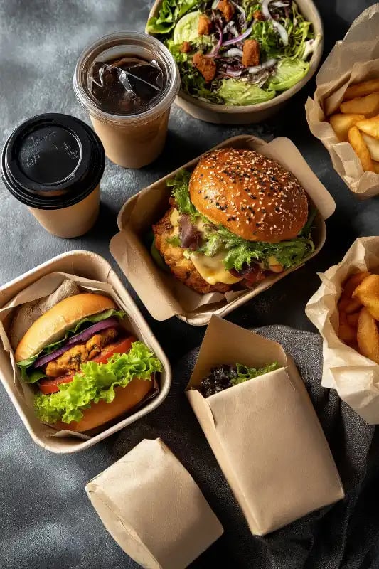 Takeout meal in kraft containers with burgers, fries, salad, and iced coffee on a dark table