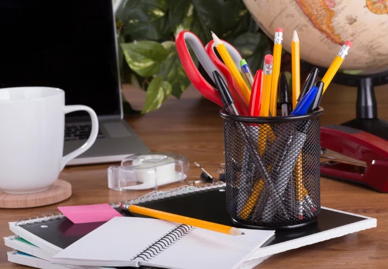 Office desk with notebook, pen cup, pencils, scissors, sticky notes, tape dispenser, and a coffee mug near a laptop