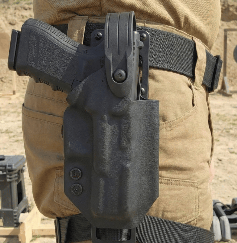 Black kydex tactical holster with a handgun secured on a shooter's belt at an outdoor gun range.