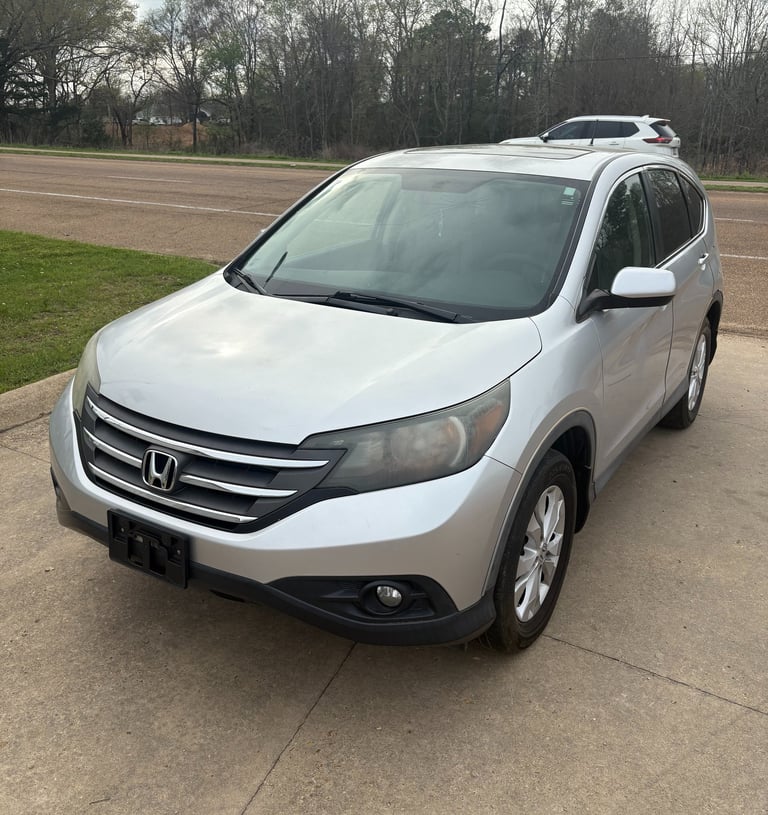 Silver Honda CR-V crossover SUV parked on a concrete driveway near a road with trees.