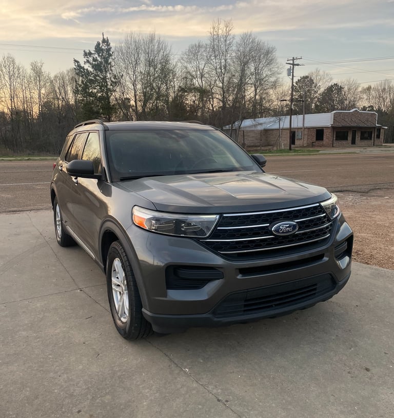 Gray Ford Explorer SUV parked on a concrete driveway during a sunset with trees in the background.
