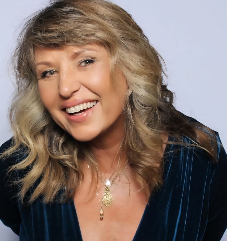 Smiling woman with wavy blonde hair wearing a blue velvet dress and gold necklaces.