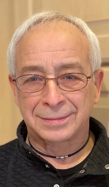 Portrait of a smiling senior man with gray hair wearing glasses and a black cord necklace.