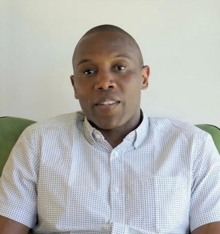 Portrait of a Black man in a patterned short-sleeve button-down shirt sitting on a green chair.