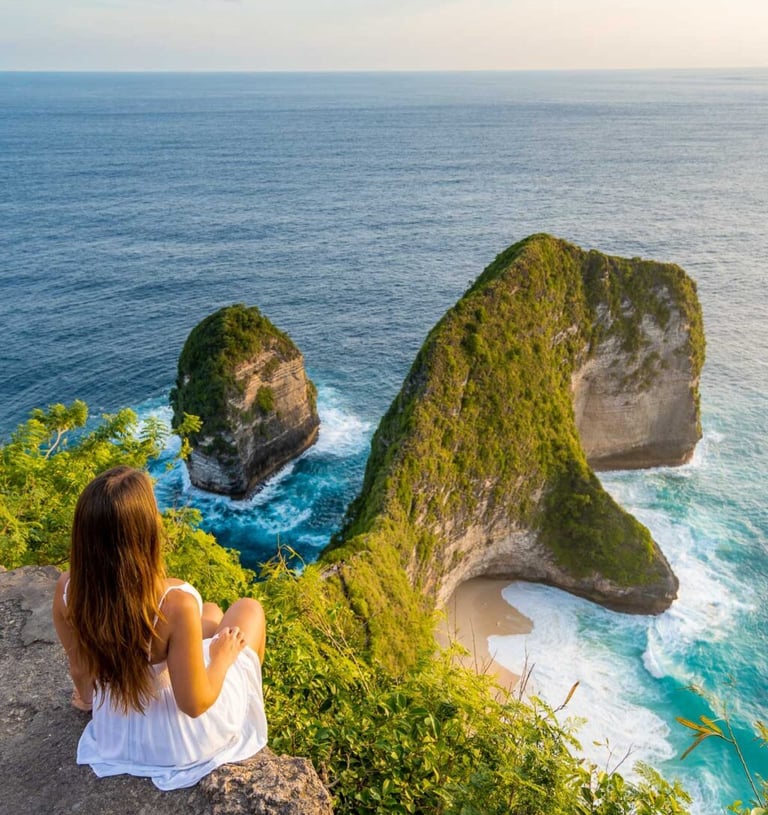 A woman in a white dress sits on a cliff overlooking Kelingking Beach in Nusa Penida, Bali.