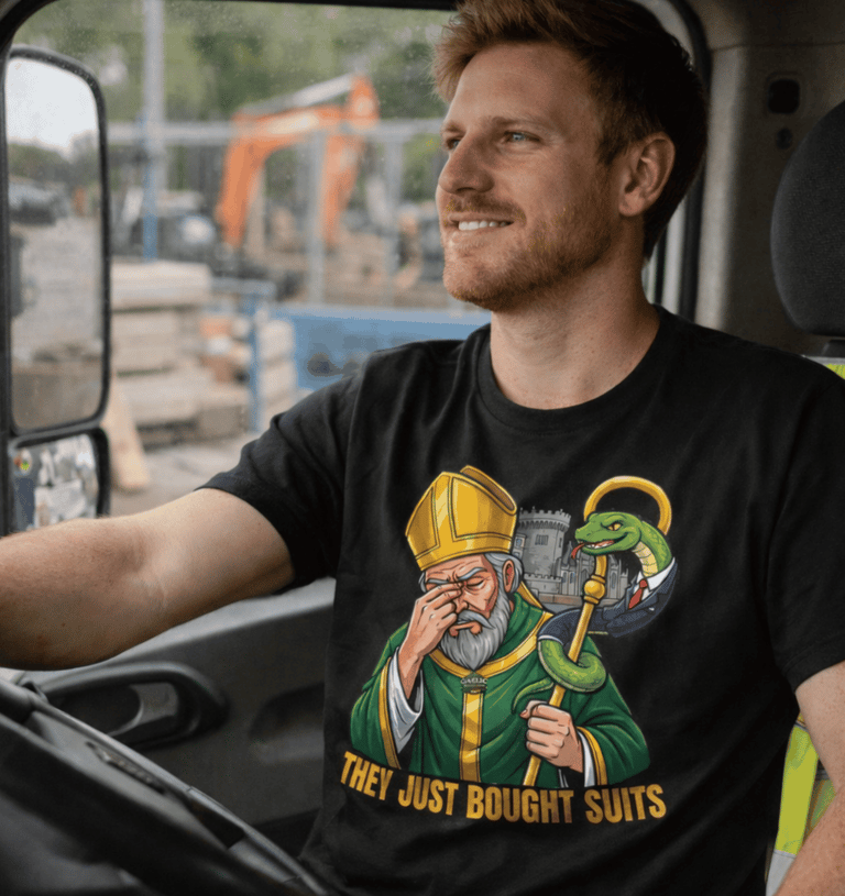 A smiling man on a couch, holding a white mug and remote, wearing a t-shirt featuring a cartoon St. Patrick looking