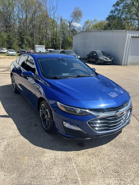 A bright blue Chevrolet Malibu sedan parked on a sunny concrete lot at a car dealership.