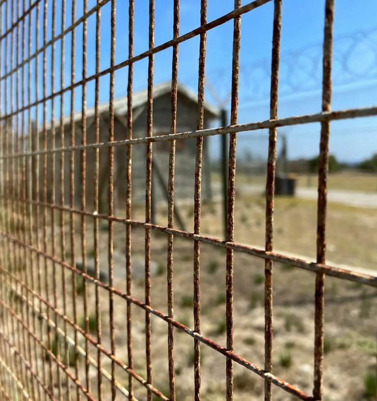 Close-up of a rusty metal wire fence with a blurred military barracks and barbed wire under a blue sky.