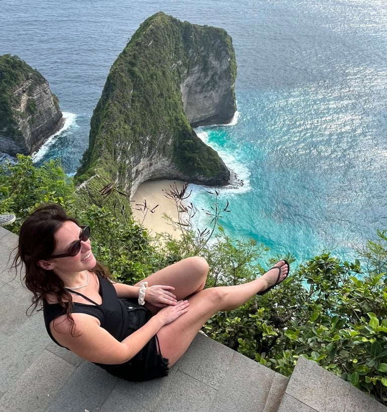 A woman sits on a cliff overlooking the iconic Kelingking Beach and T-Rex Bay in Nusa Penida, Bali.