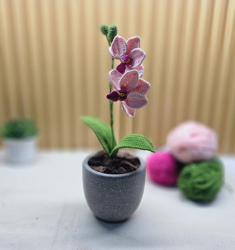 Handmade pink crochet orchid plant in a grey pot with green leaves and yarn balls in the background.
