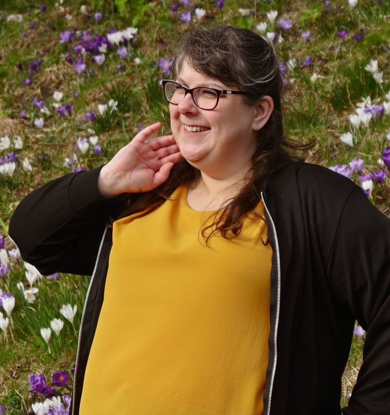 Laura is standing in front of a field of purple and white flowers, looking away from the camera with a soft smile on her face