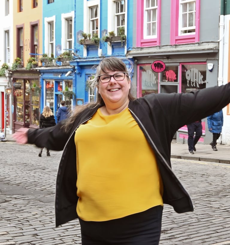Laura is dancing with her arms out on a cobbled a street with brightly coloured buildings behind her