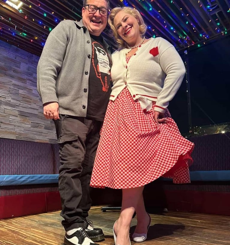 A smiling couple in retro-style clothing poses under colorful lights at a rockabilly event.