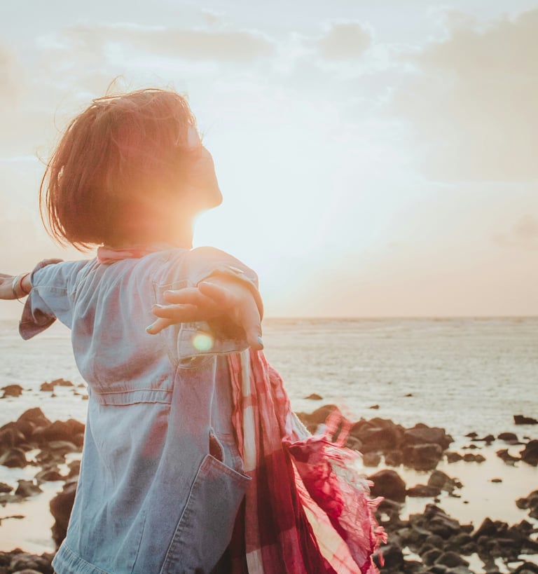 une femme à la retraite, debout, détendue, bras ouverts au bord de la mer, qui inspire le bonheur devant le coucher de soleil