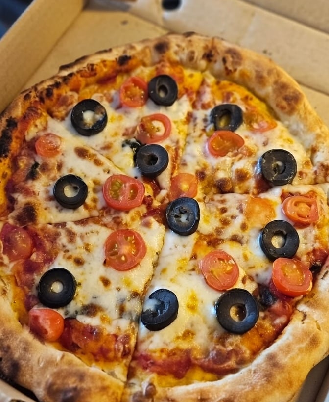 Freshly baked pizza with melted mozzarella cheese, black olives, and cherry tomatoes in a cardboard box.