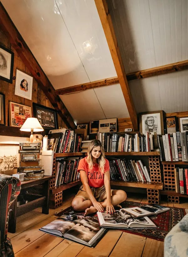 A woman sits on the floor of a cozy attic home library reading books near wooden shelves.