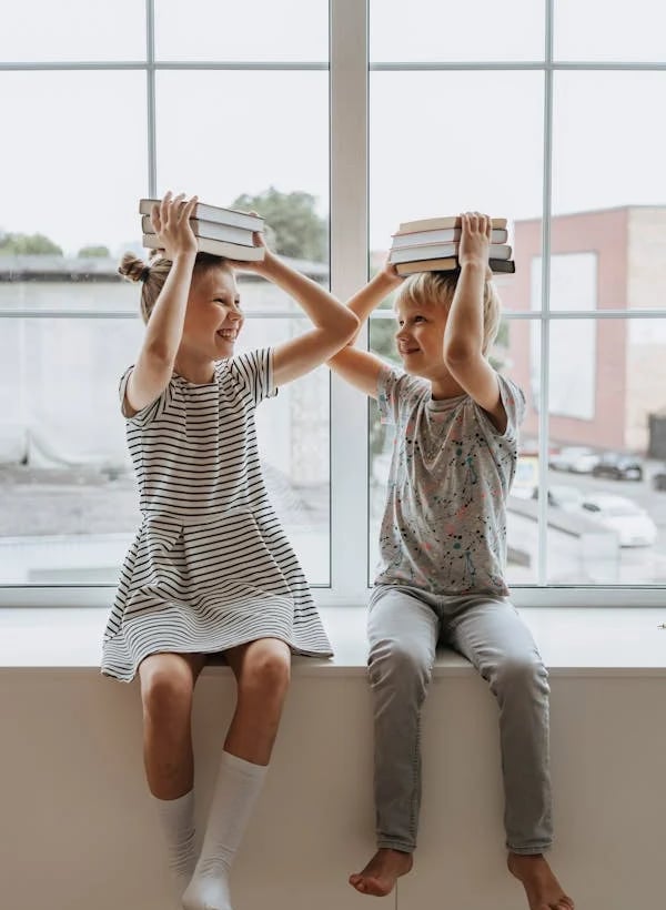 Du laimingi vaikai sėdi ant palangės ir laiko knygas ant savo galvų.