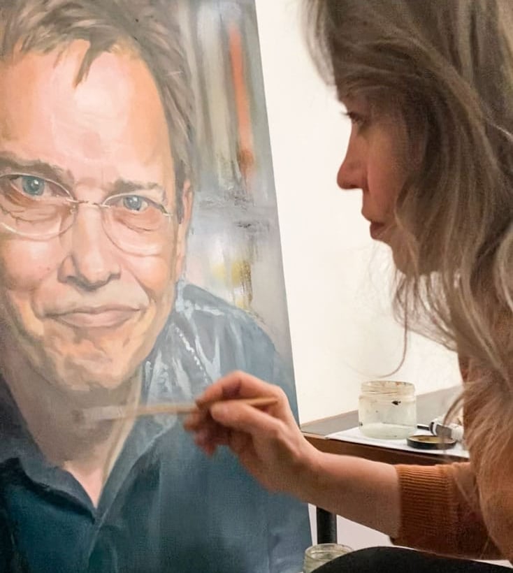 Stefanie placing final touches on oil portrait painting of smiling man with glasses.