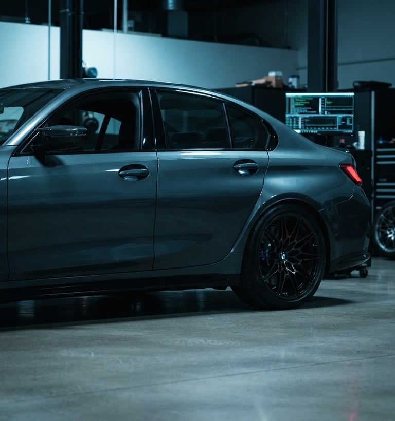 Low-angle profile of a dark grey BMW M-series parked in a clean, modern workshop bay, cool blue ambient light highlighting its metallic paint, with a blurred diagnostic screen displaying code in the background. High-contrast studio lighting.