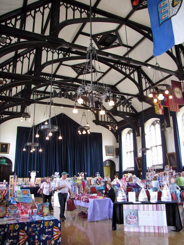 Indoor craft fair in a gothic hall with vaulted ceilings, hanging chandeliers, and vendor displays.