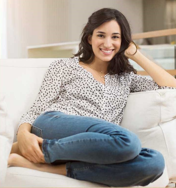A smiling woman in a patterned blouse and jeans sits comfortably on a white sofa.