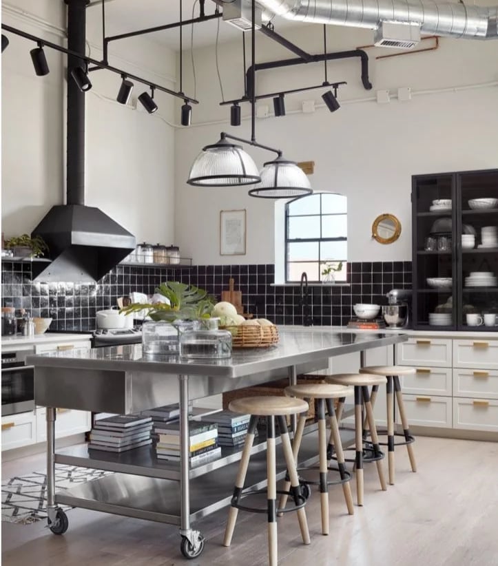 A large stainless steel table on wheels used as a kitchen island with bar stools and a lower shelf for cookbooks.