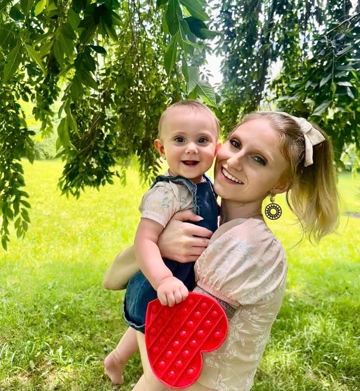 Mother holding baby close under a tree, both smiling at the camera, baby holding heart-shaped toy.
