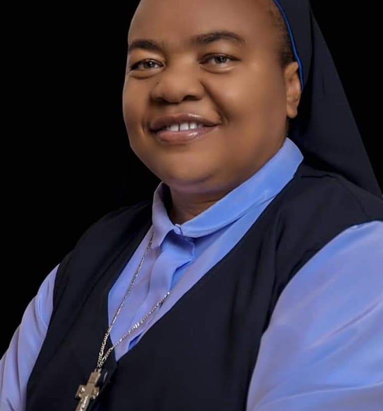 Smiling Catholic nun wearing a blue habit and silver cross necklace against a black background.