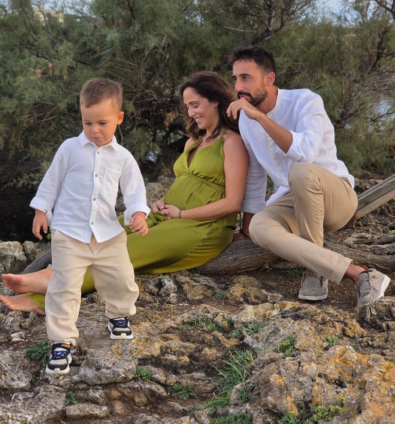 A family of three sitting on rocky terrain near a river with greenery in the background, enjoying an outdoor moment.