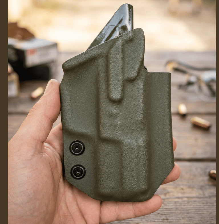 Hand holding an olive drab Kydex IWB holster for concealed carry over a wooden workbench with ammo.