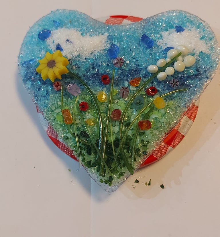Handmade heart-shaped fused glass art featuring a floral meadow with a yellow sunflower and blue sky.