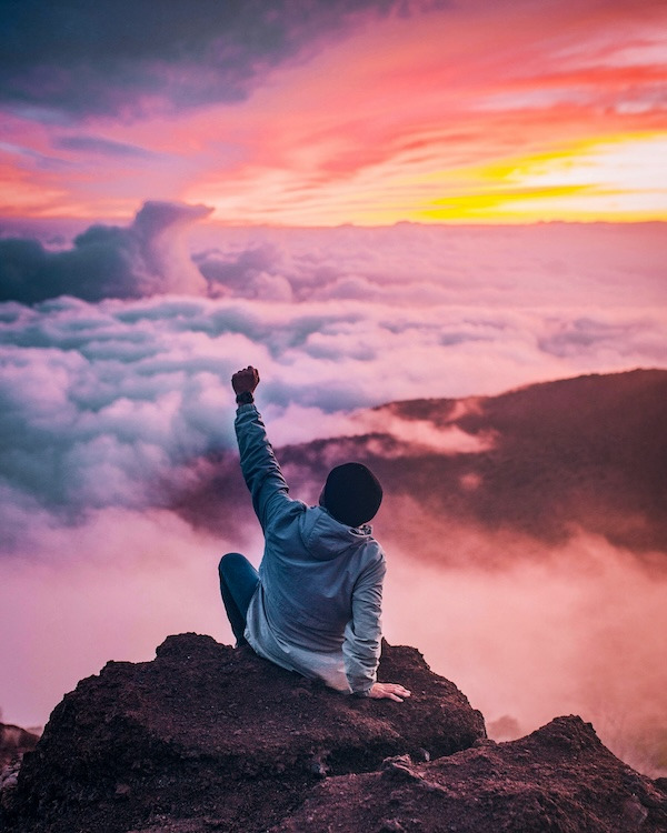 Young man on top of a mountain, above the clouds, with the sun setting in the background.