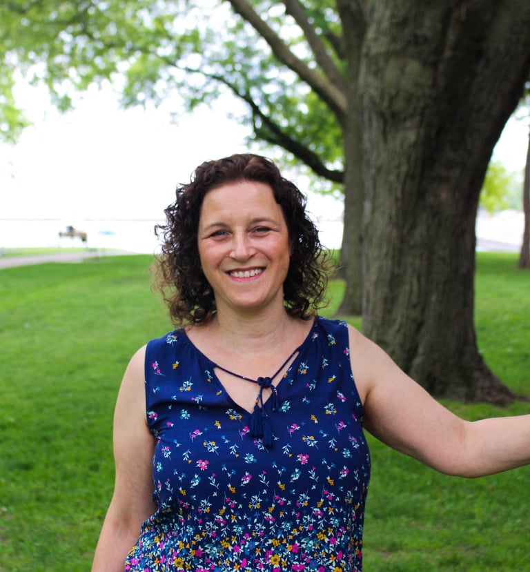 Picture of Natanya in a park, hand on a tree, water in the background.