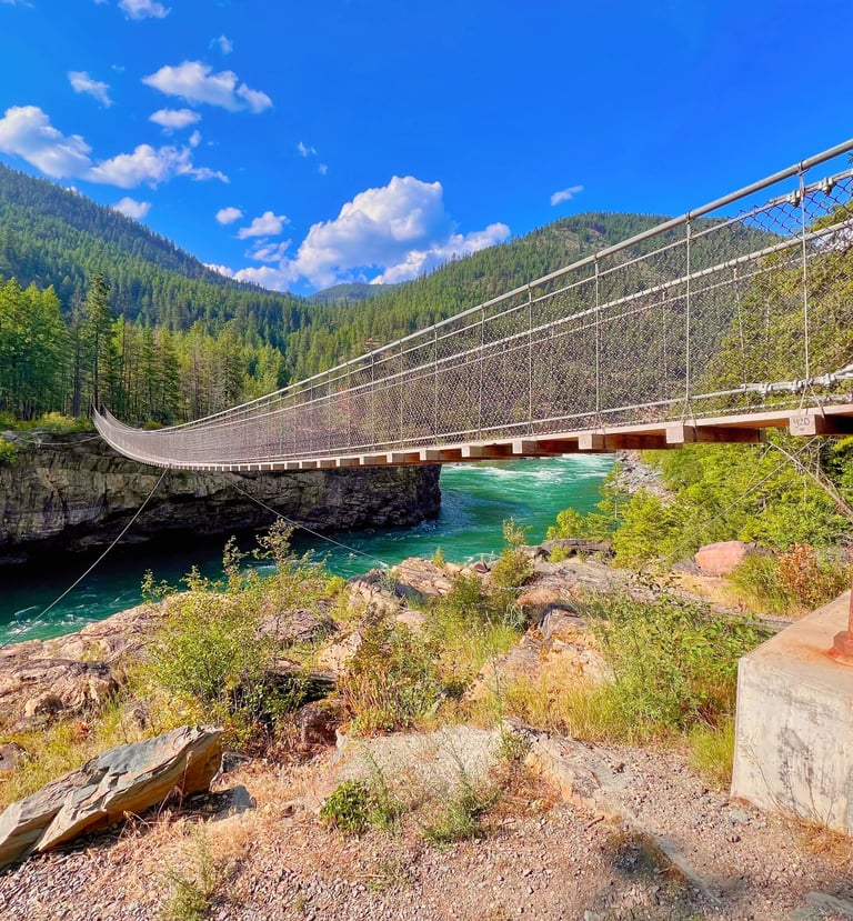 A long suspension footbridge spanning a turquoise river in a mountain canyon under a bright blue sky.